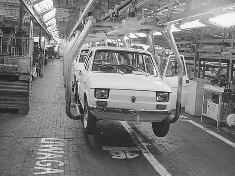 Black-and-white photograph of a small white car carried on a hanging cradle along a brick-floored factory aisle. Opens a larger view