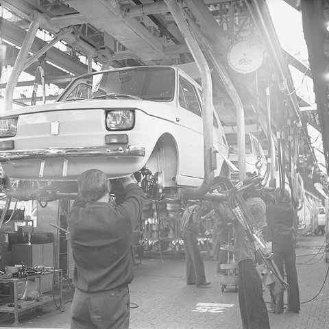 Black-and-white photograph of a small car hanging on an overhead conveyor while workers reach up to fit parts beneath it. Opens a larger view