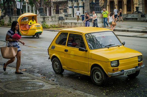 Yellow small two-door car on a wet road as a woman with a handbag walks past and a yellow coco-taxi waits behind.