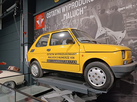 Yellow small two-door car on a raised lift with black lettering on its door, a large black-and-white factory mural behind. Opens a larger view