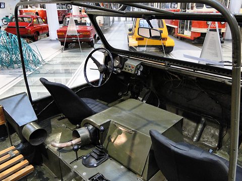 Open olive-green cockpit with two black seats, a black steering wheel and a gear lever on a raised tunnel. Opens a larger view