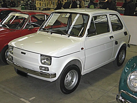 Small white two-door car with chrome bumpers in an exhibition hall between red cars, visitors behind. Opens a larger view