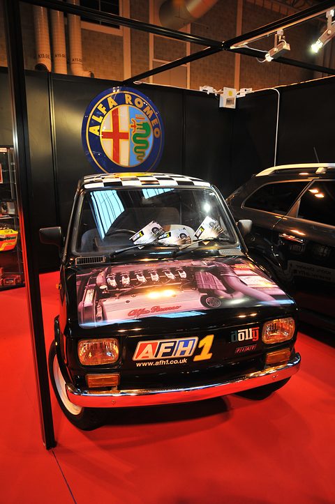 Black small car with a chequered roof and an engine picture printed on its bonnet on a red-carpeted show stand. Opens a larger view