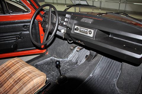 Black dashboard with a radio, a thin black steering wheel and a floor gear lever beside a brown checked seat. Opens a larger view