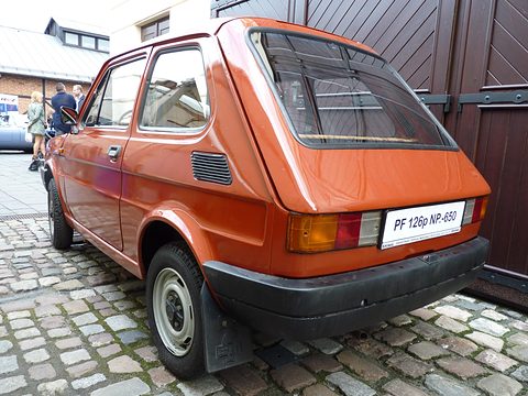 Rear three-quarter of an orange small hatchback with a large rear window and black bumper on a cobbled yard. Opens a larger view