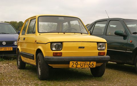 Yellow small two-door car with black plastic bumpers and side strips parked on gravel between two modern cars. Opens a larger view