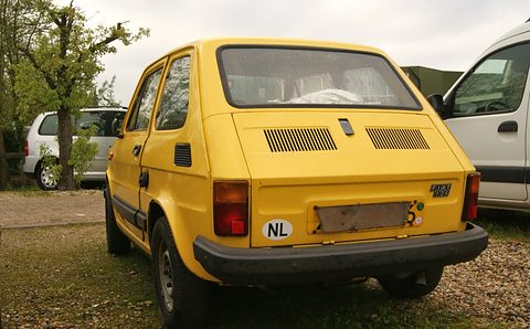 Rear three-quarter of a yellow small car with black bumpers, slotted engine lid and an NL sticker on gravel. Opens a larger view