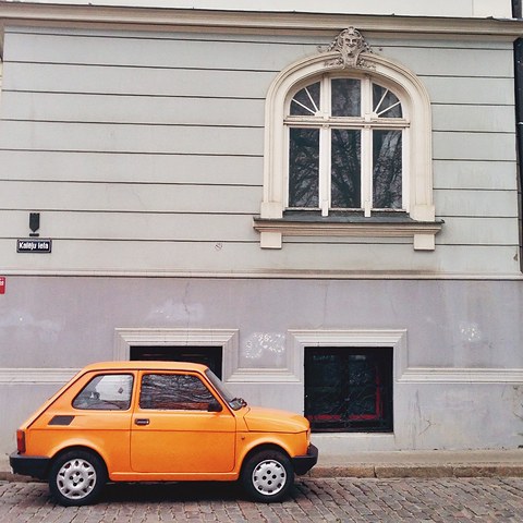 Orange small two-door car with black bumpers parked on cobbles beneath a grey building with an arched window. Opens a larger view