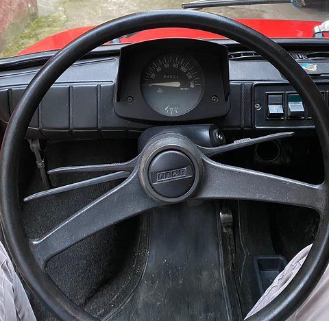 Black three-spoke steering wheel with a FIAT horn button in front of a round speedometer and rocker switches. Opens a larger view
