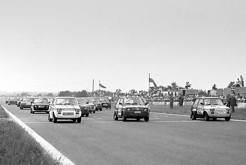 A pack of small white two-door saloons with race numbers coming down a wide circuit straight, spectators beyond.