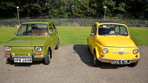 A boxy green small car and a rounded yellow small car parked side by side on tarmac in front of a grass bank.