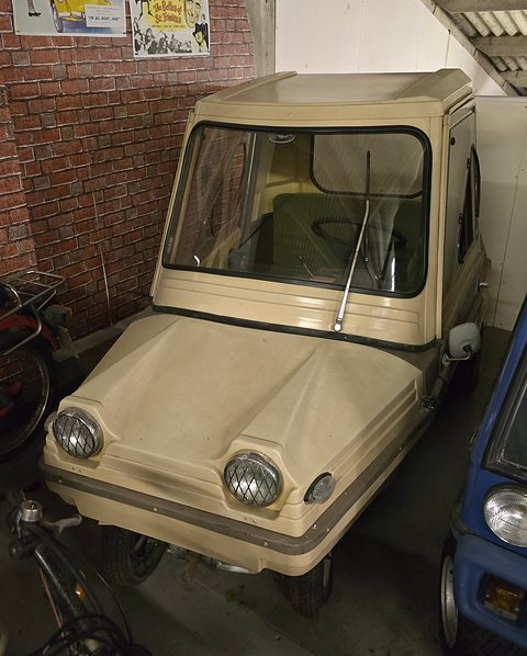 Beige boxy two-seater with round headlamps in grilled pods and a raked bonnet, by a brick wall with film posters. Opens a larger view