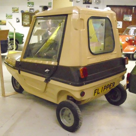 Beige and brown boxy car from behind and to the right, with large acrylic side windows and a FLIPPER plate. Opens a larger view