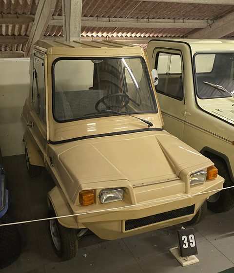 Beige boxy two-seater with square headlamps and orange indicators under a corrugated roof, a rope and exhibit number 39 in front.