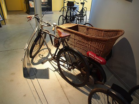 An old bicycle with a large wicker basket on the side, a red motor-wheel tank by its rear wheel, in a sunlit museum corridor. Opens a larger view