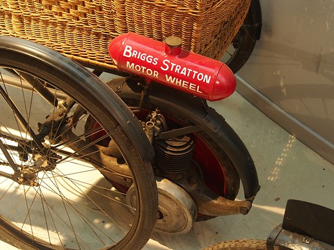 A red tank lettered Briggs Stratton Motor Wheel over a small engine in a spoked wheel, beside a bicycle wheel and wicker basket. Opens a larger view