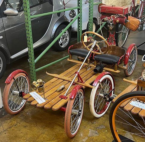 A slatted wooden buckboard on four red bicycle wheels with two small seats, a long steering column and a brass lamp. Opens a larger view