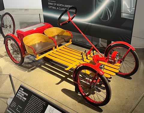 A red electric buckboard with slatted wooden deck, two wooden bucket seats and a long steering column, beside its museum label. Opens a larger view