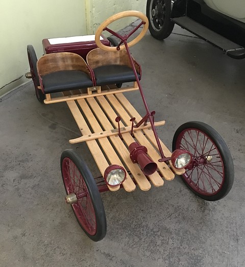 A slatted wooden buckboard with dark red wire wheels, two black seats and twin headlamps, seen from above the front. Opens a larger view