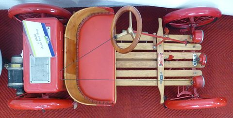 Overhead view of a red buckboard: battery box with a placard, padded red seat, wooden steering wheel and slatted deck. Opens a larger view
