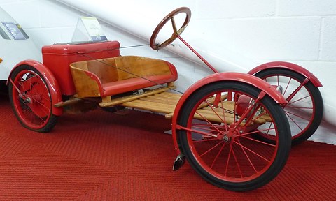 A red-wheeled wooden buckboard with a wooden seat and red battery box on a red carpet against a white block wall. Opens a larger view