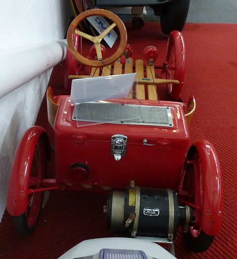 Looking down on the back of a red buckboard: a red battery box with a badge, and a black and brass electric motor below it.