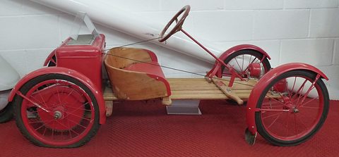 Side view of a small red buckboard car with a wooden seat, slatted deck and long steering column. Opens a larger view