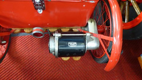 A black cylindrical electric motor with a small maker’s plate beneath a red box, beside a red spoked wheel. Opens a larger view