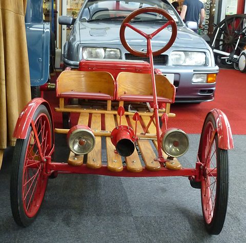 Front view of a slatted wooden buckboard with two lamps and a horn on the front bar, red wheels and a steering wheel. Opens a larger view