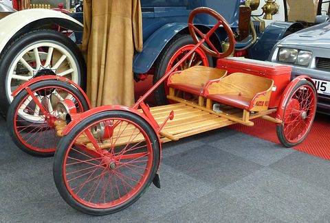 A red-wheeled slatted buckboard with twin wooden seats and a red box, parked before larger veteran cars and a hanging coat. Opens a larger view
