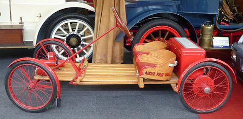 Side view of a small red-wheeled wooden buckboard with two seats and a box at the back, veteran cars behind. Opens a larger view