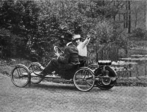 A driver and a waving passenger on a flat wooden buckboard with four bicycle wheels and an engine-driven fifth wheel behind.