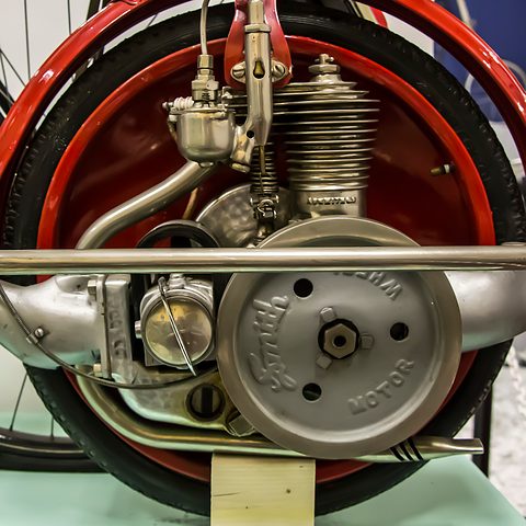 Close view of a small finned single-cylinder engine with carburettor and a cover lettered Smith Motor inside a red-rimmed wheel.