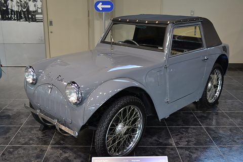 Grey small car with a dark fabric top, round headlamps on the wings and chrome wire wheels on a dark tiled floor. Opens a larger view