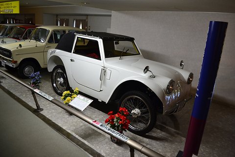 A small white two-seater with a black top and wire wheels in a line of older cars, a blue pillar beside it. Opens a larger view