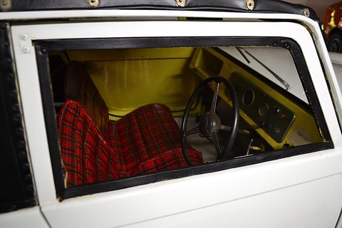 View through a car door of red tartan seat cushions, a thin black steering wheel and a yellow-painted dashboard.