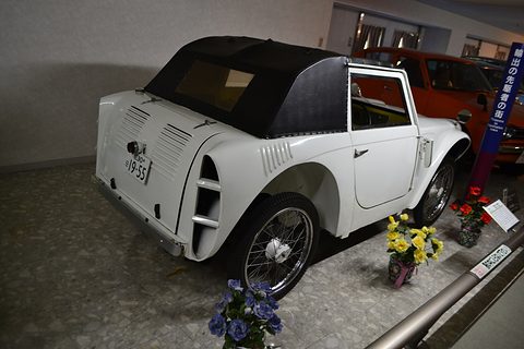 Rear three-quarter view of a small white car with a black fabric top, louvred tail panel and wire wheels in a museum row.