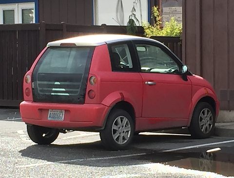 Red tall two-door city car with a large black glass tailgate and grey plastic bumpers in a car park. Opens a larger view