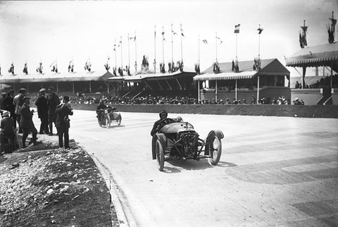 A small open racing cyclecar with an exposed engine at its nose on a curving concrete track, a sidecar outfit and spectators behind. Opens a larger view