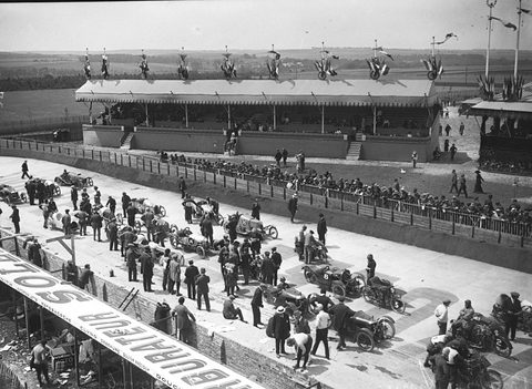 High view over rows of small racing cars and men on a pale apron, long grandstands and open fields behind. Opens a larger view