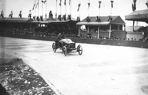 A small dark racing cyclecar with two men aboard on a pale road, grandstands with flags behind. Opens a larger view