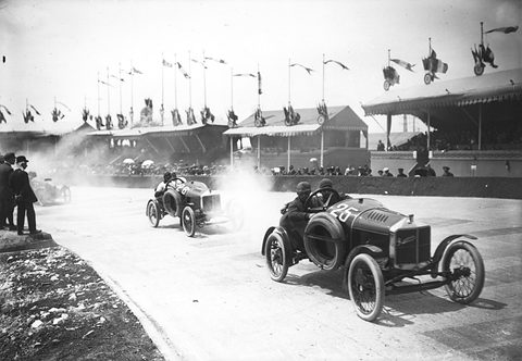 Two racing cyclecars throw up dust on a pale road, the nearer numbered 26, grandstands and flags behind. Opens a larger view