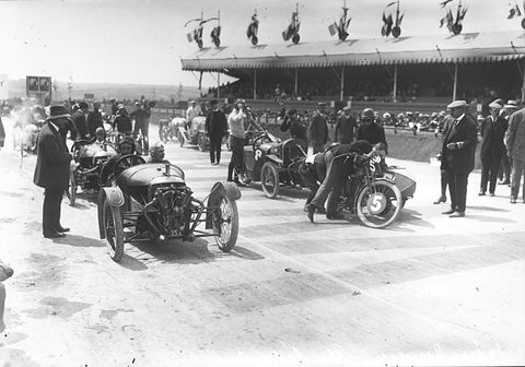 A small racing car with an exposed engine pulls away from a crowd of officials and motorcycle sidecars, grandstand behind. Opens a larger view