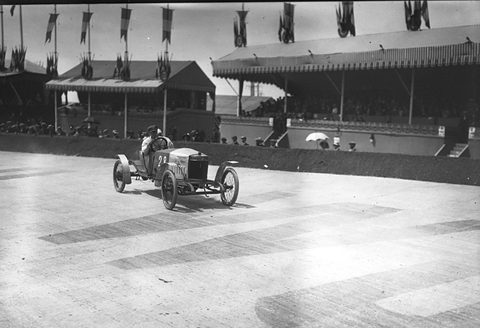A small open racing cyclecar on a wide pale road before long flag-decked grandstands full of spectators. Opens a larger view