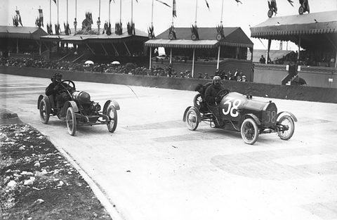 Two small racing cyclecars with goggled drivers on a pale road curve, flag-hung grandstands behind.