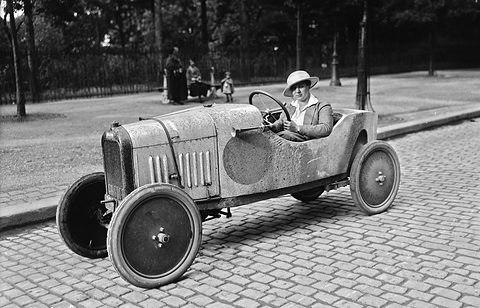 A woman in a white hat at the wheel of a small pale cyclecar with disc wheels on a cobbled street, trees and railings behind. Opens a larger view