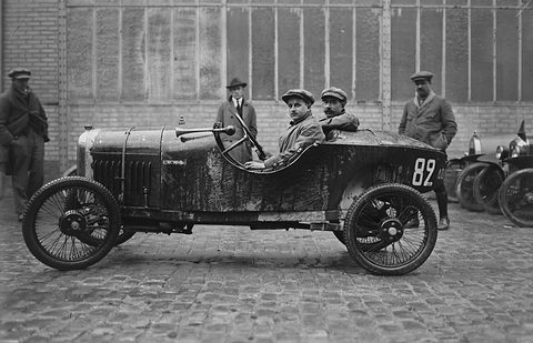 Two men in caps in a dark muddy cyclecar numbered 82 on cobbles before a glazed hangar, others standing by. Opens a larger view