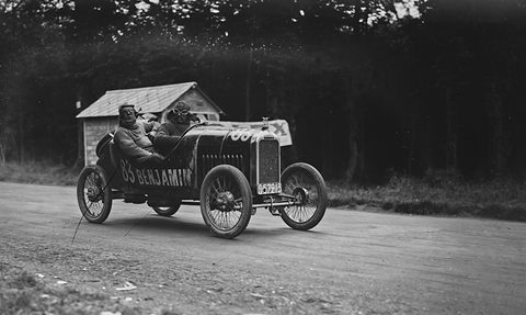 A dark cyclecar lettered Benjamin with two men aboard rounds a bend before a small hut and dark trees. Opens a larger view