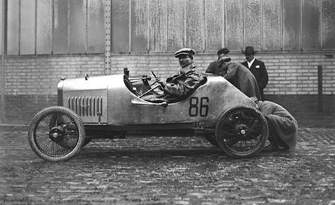 A driver in a pale pointed-tail racing cyclecar numbered 86 on cobbles, a mechanic bending at its rear, a hangar behind. Opens a larger view