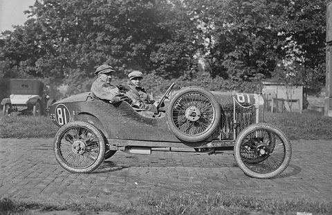 Side view of a small cyclecar with a spare wheel on its flank and two people aboard on cobbles before trees. Opens a larger view